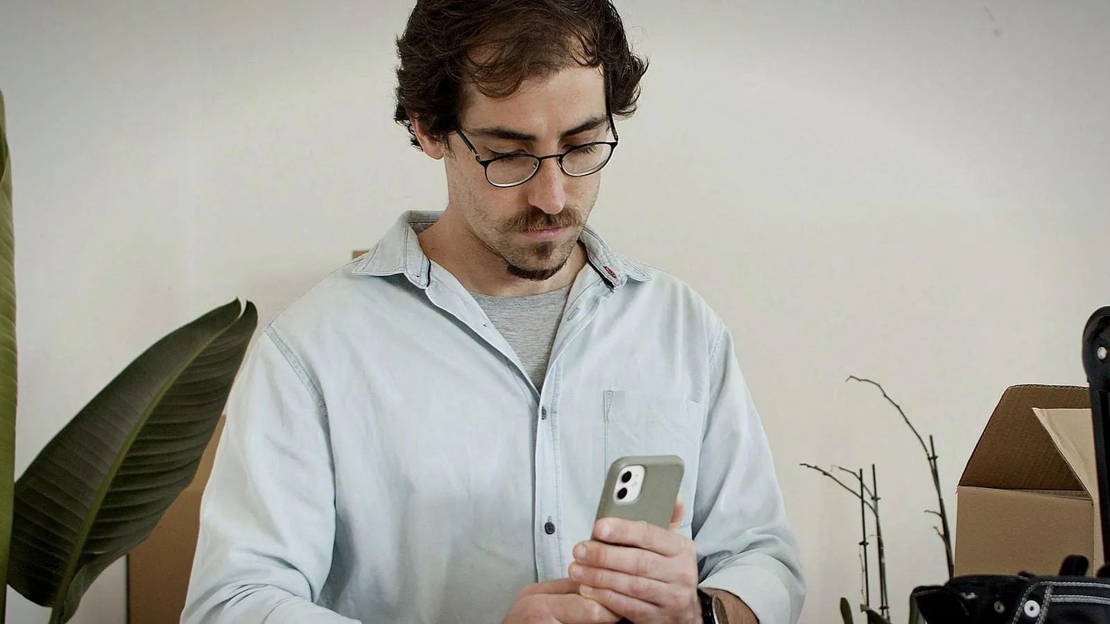 A young man in a small sneaker studio, frowning at his phone beside a box and a shoe on the bench.