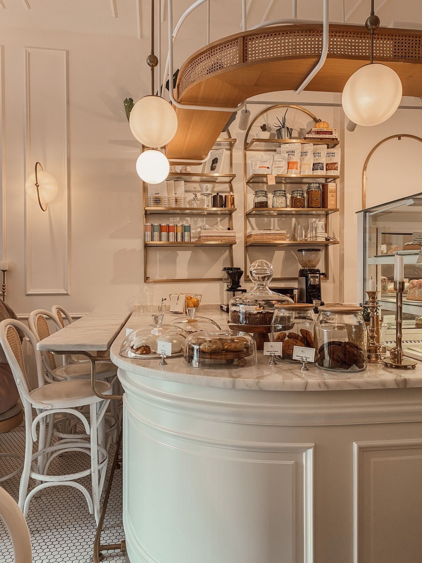 A café counter with nobody at it: cakes under glass domes on white marble, jars on brass shelves behind.