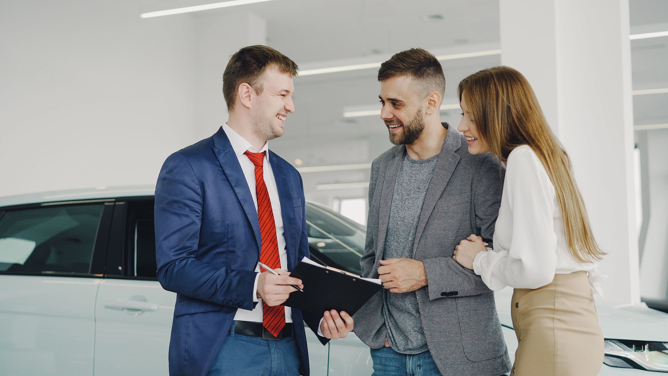 A car salesman in a red tie chatting with a smiling couple in a bright showroom.