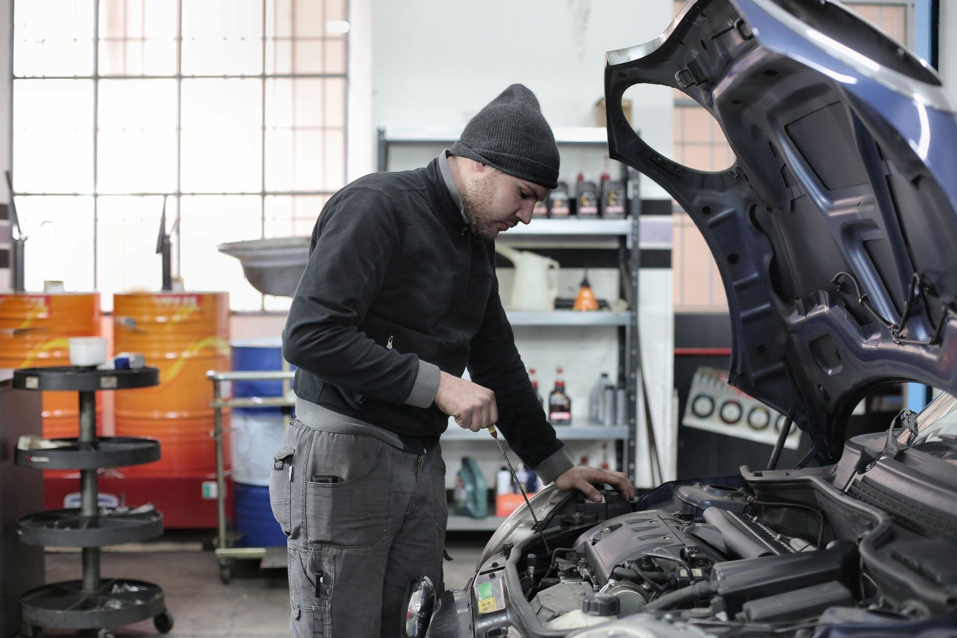 A mechanic in a beanie working on the engine under a car’s open hood in his workshop.