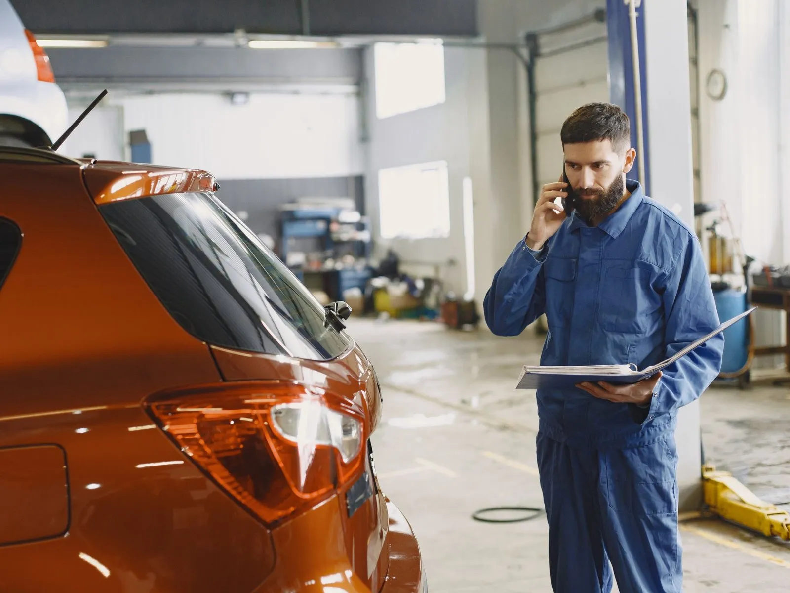 A service technician on the phone with a folder of paperwork, beside a customer car in the service bay.
