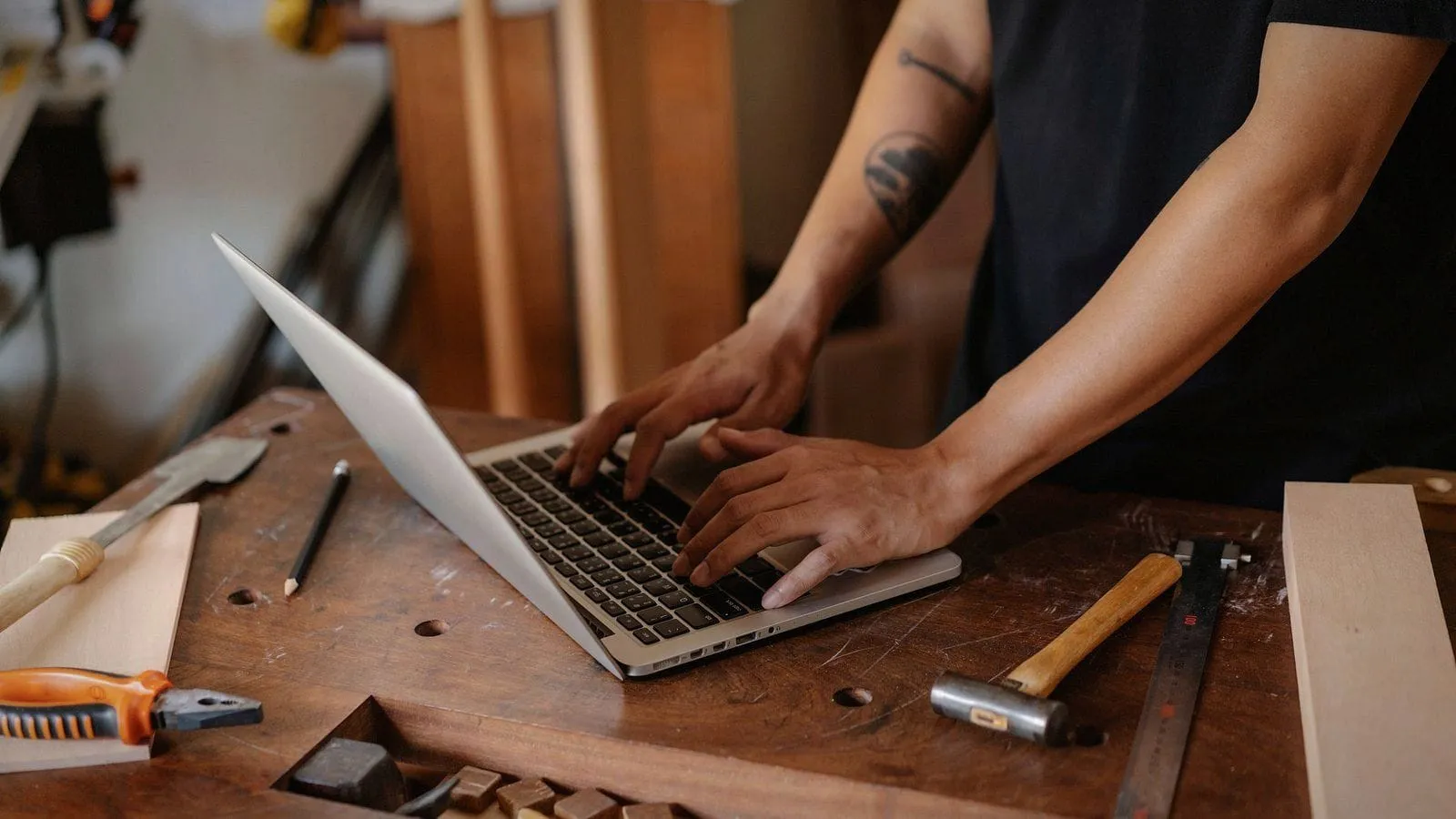 A woodworker typing on a laptop at his workbench, a hammer and tools beside it.