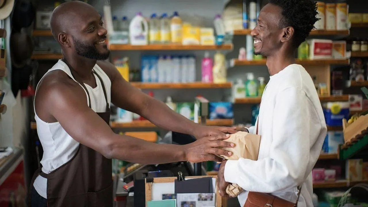A shopkeeper smiling as he hands a paper bag to a customer.