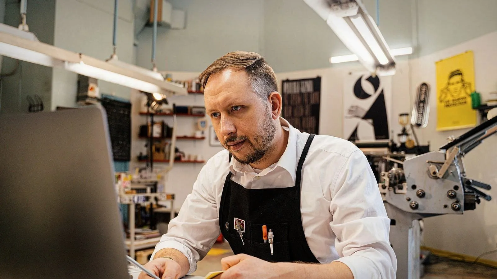 A print-shop owner in an apron at his desk, notes in hand, looking up thoughtfully.