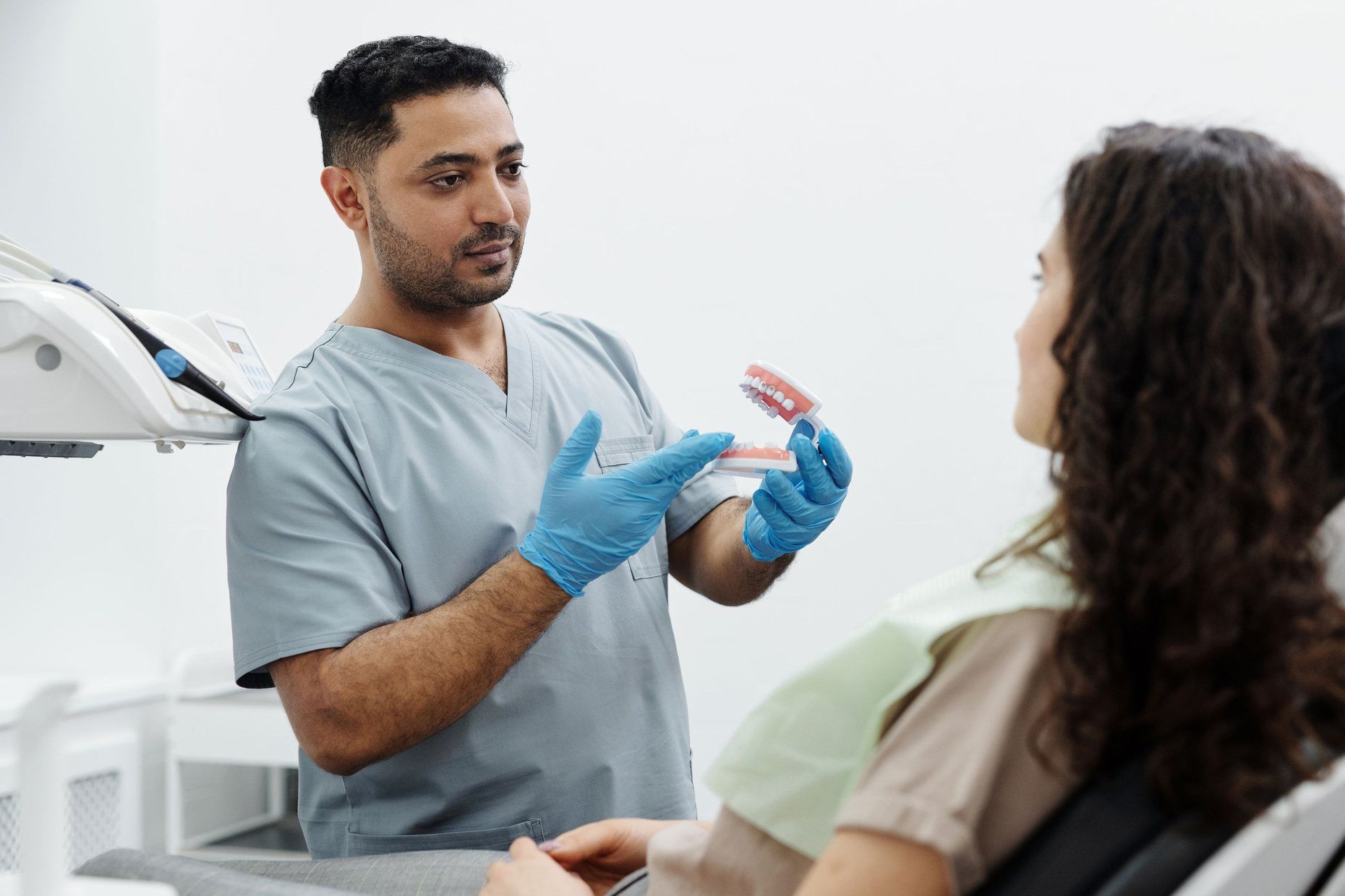 A dentist in scrubs explaining a treatment to a seated patient with a model of the teeth.