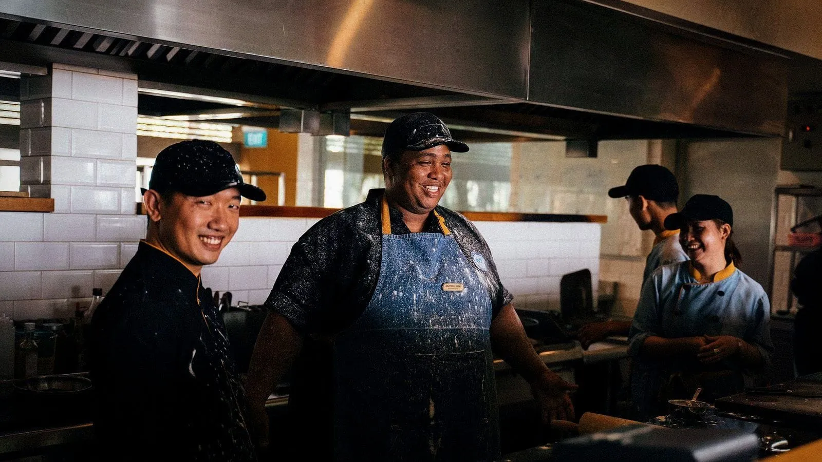 A restaurant kitchen crew in caps and aprons laughing together at the pass.