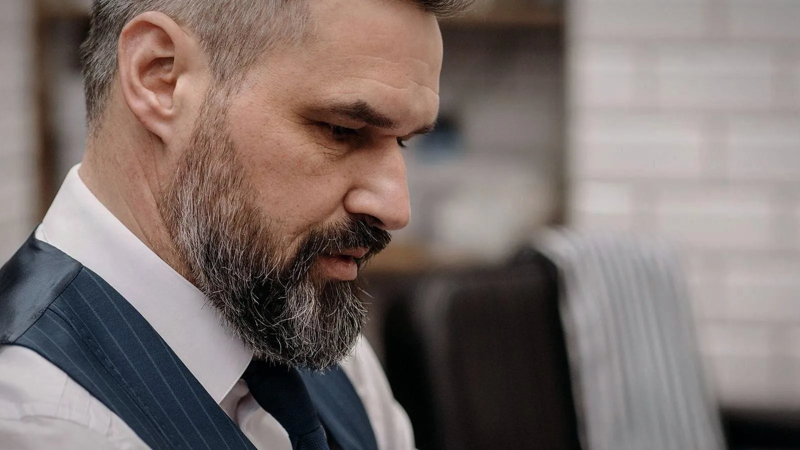A barber in a waistcoat in his shop, frowning down at a message on his phone.