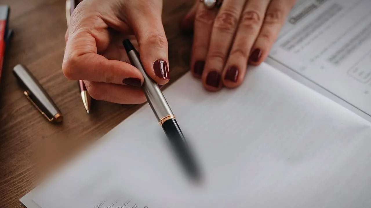 A hand holding a pen over printed papers on a desk.