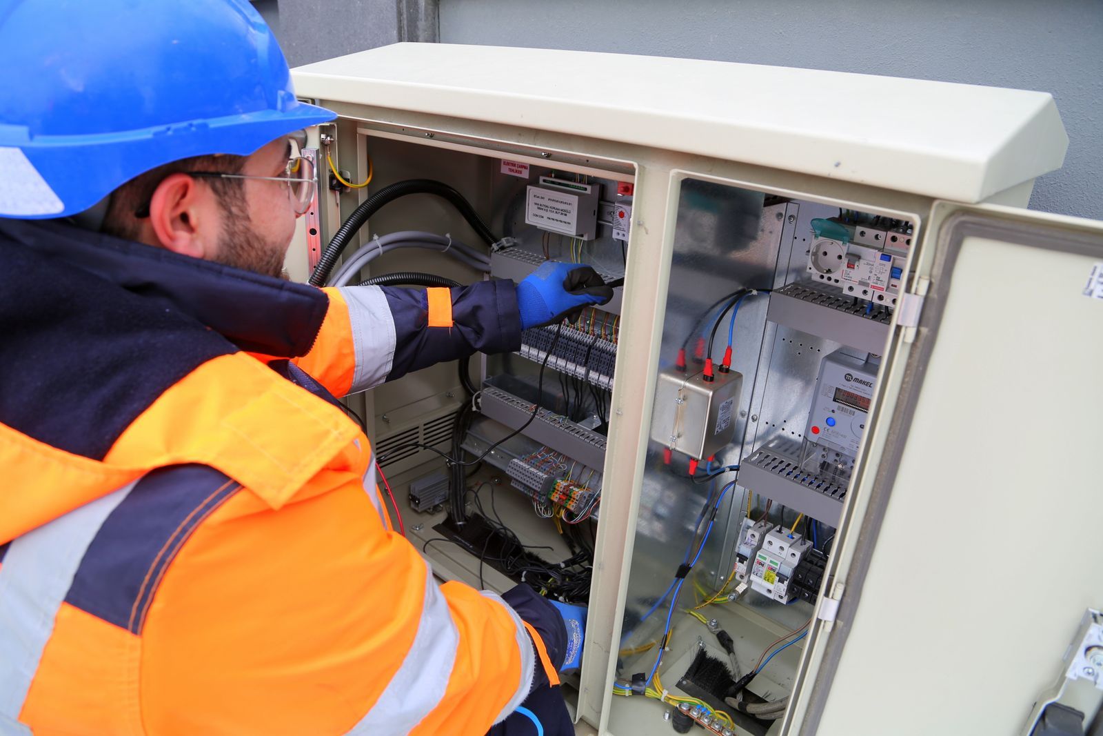 A technician in a hard hat and high-visibility vest working on an open electrical cabinet.