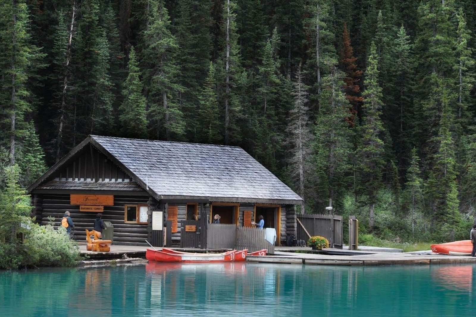 A wooden lodge on the edge of a mountain lake, canoes tied up at the dock and forest behind.