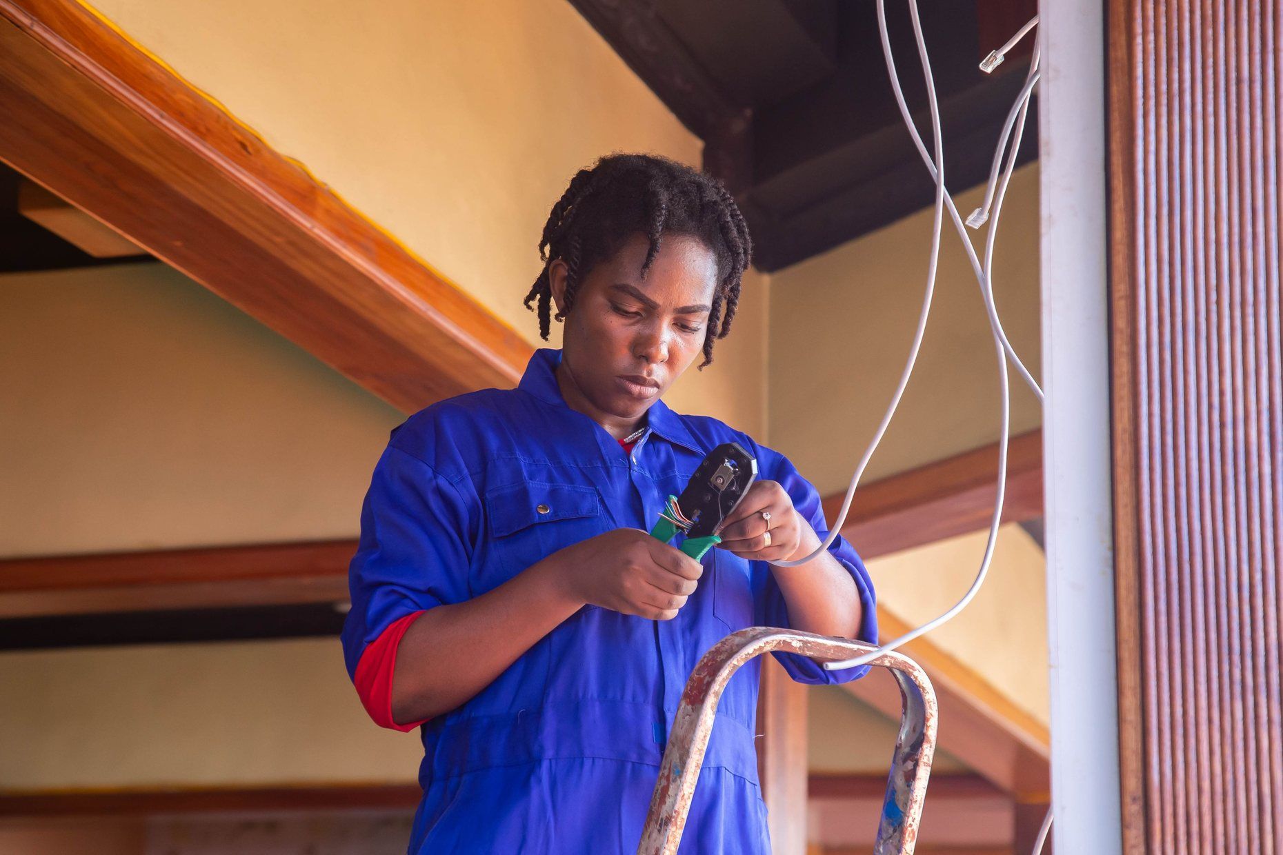 An electrician in blue overalls on a stepladder, stripping the end of a new cable.