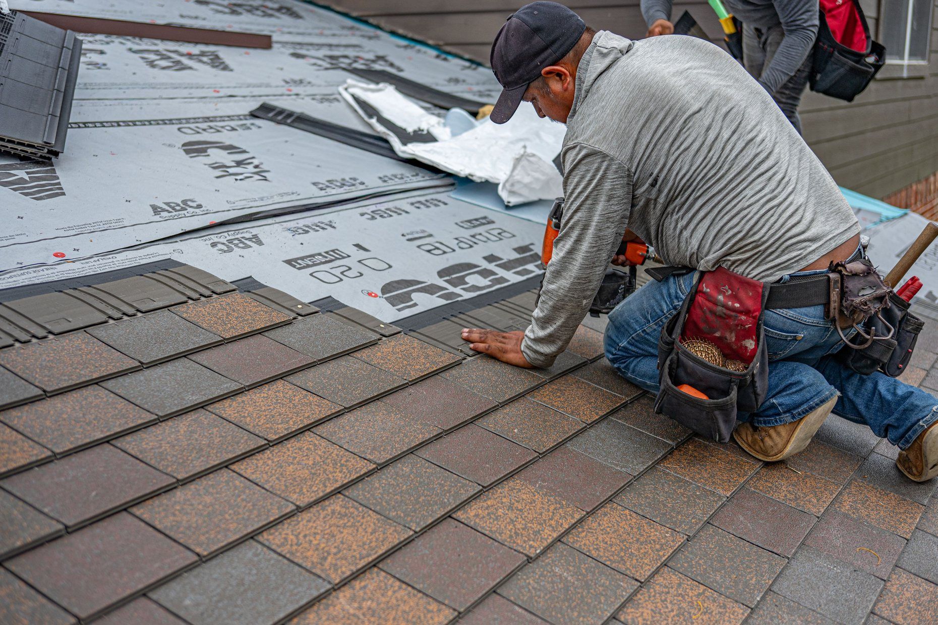 A roofer kneeling on a suburban roof, laying a new row of asphalt shingles.