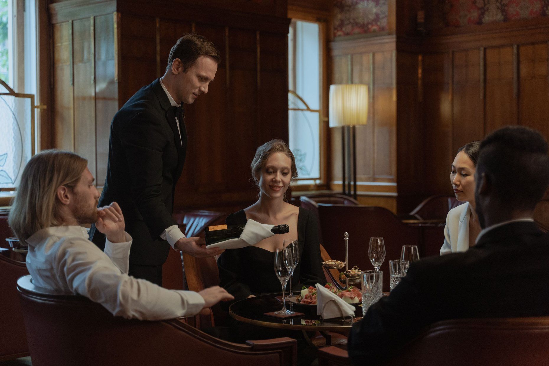 A waiter in a dinner jacket serving guests at a table in a wood-panelled restaurant.