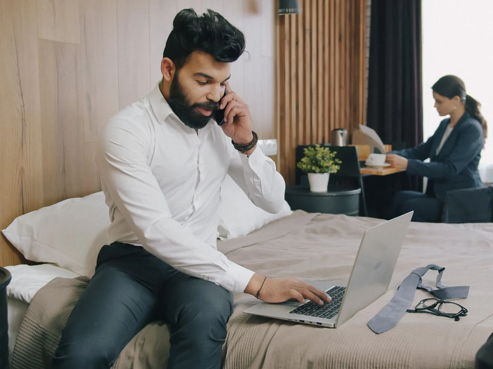 A traveler on the phone on a hotel bed, his laptop open beside him.