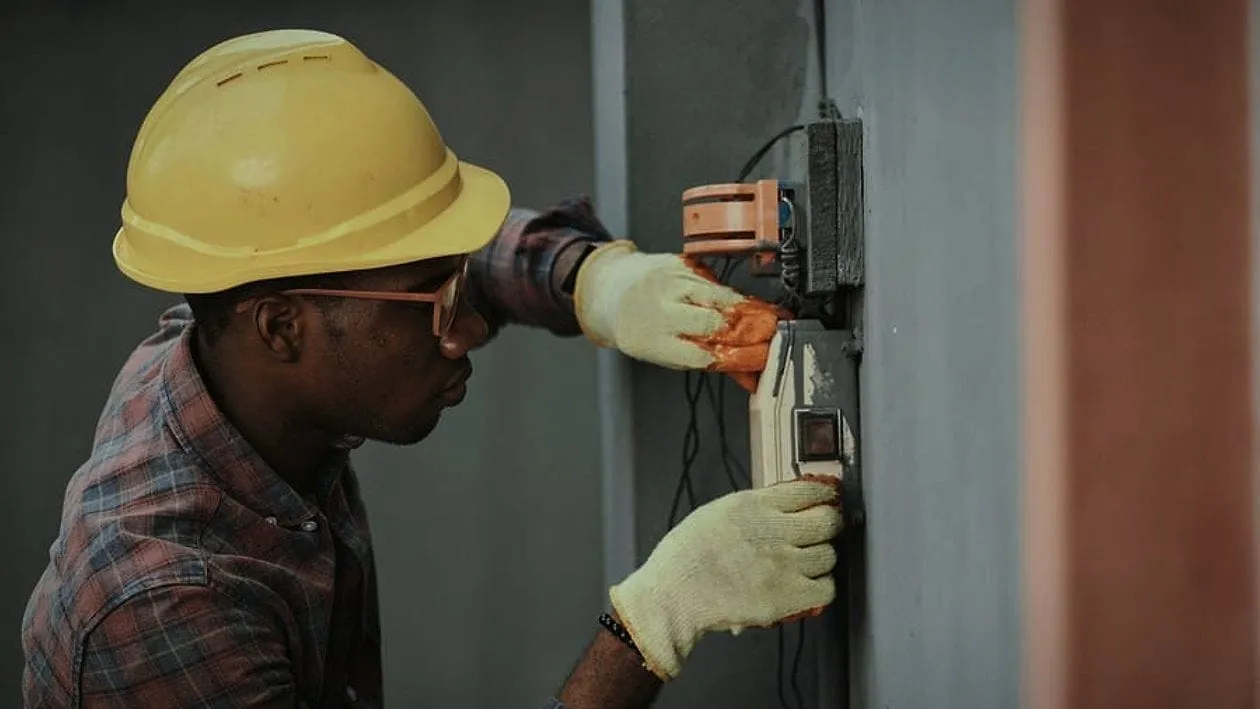 A tradesman in a hard hat and gloves working on a meter on a wall.