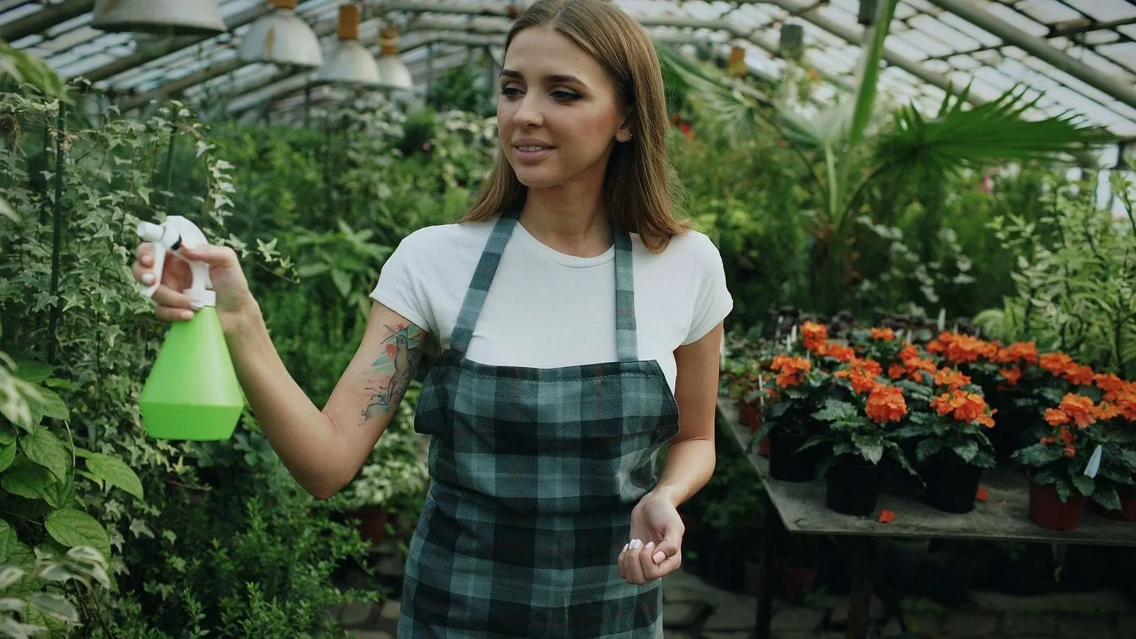 A woman in an apron misting plants in a sunlit greenhouse full of flowers.