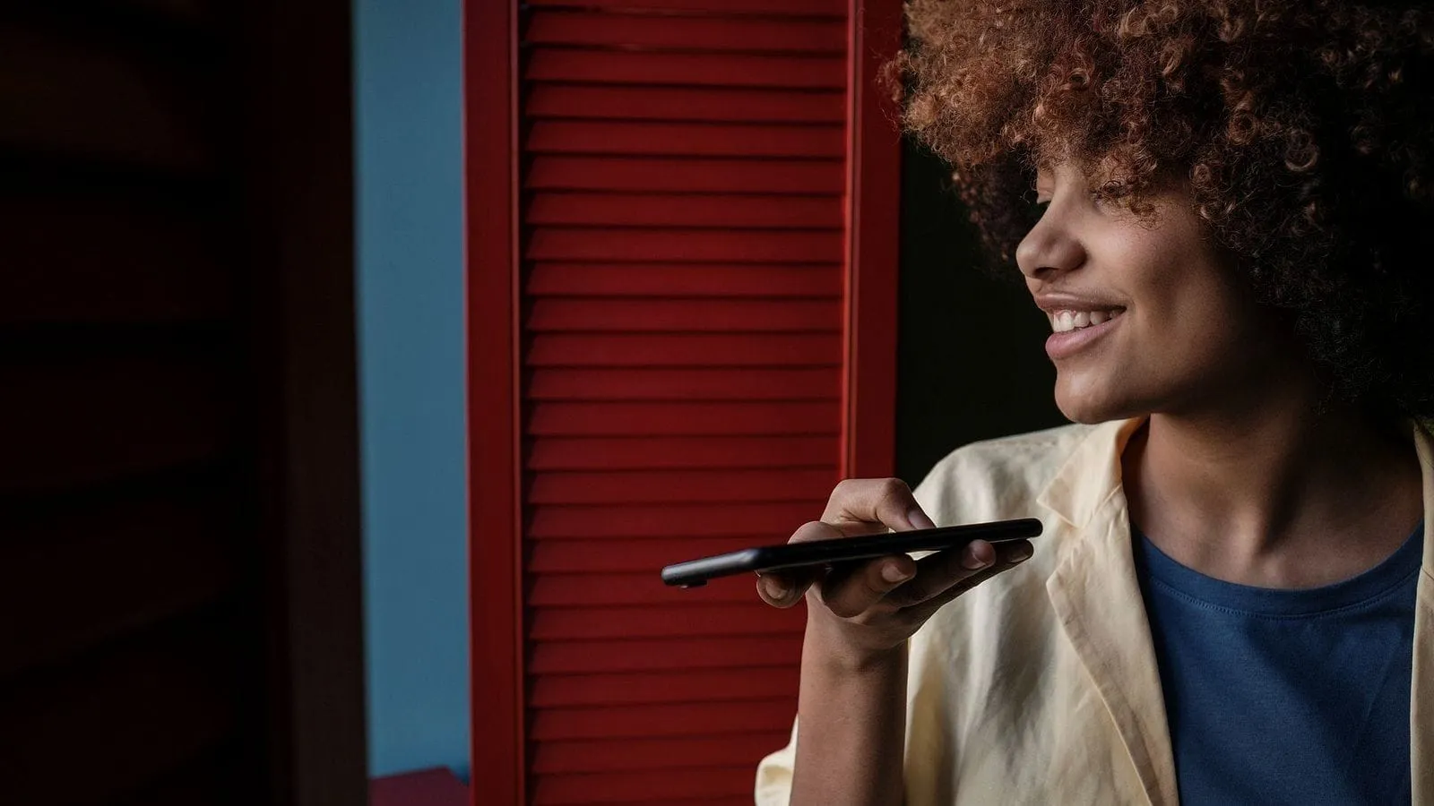 A smiling woman outside a shop with red shutters, speaking a question into her phone.