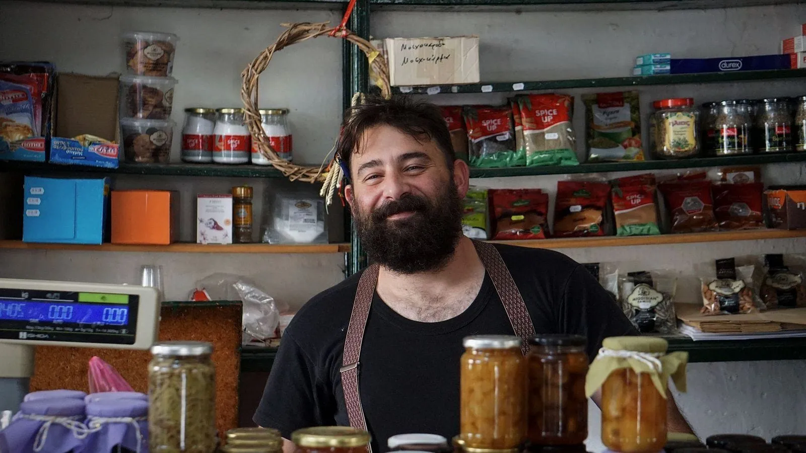 A bearded grocer smiling behind his counter of jars, shelves of goods behind him.