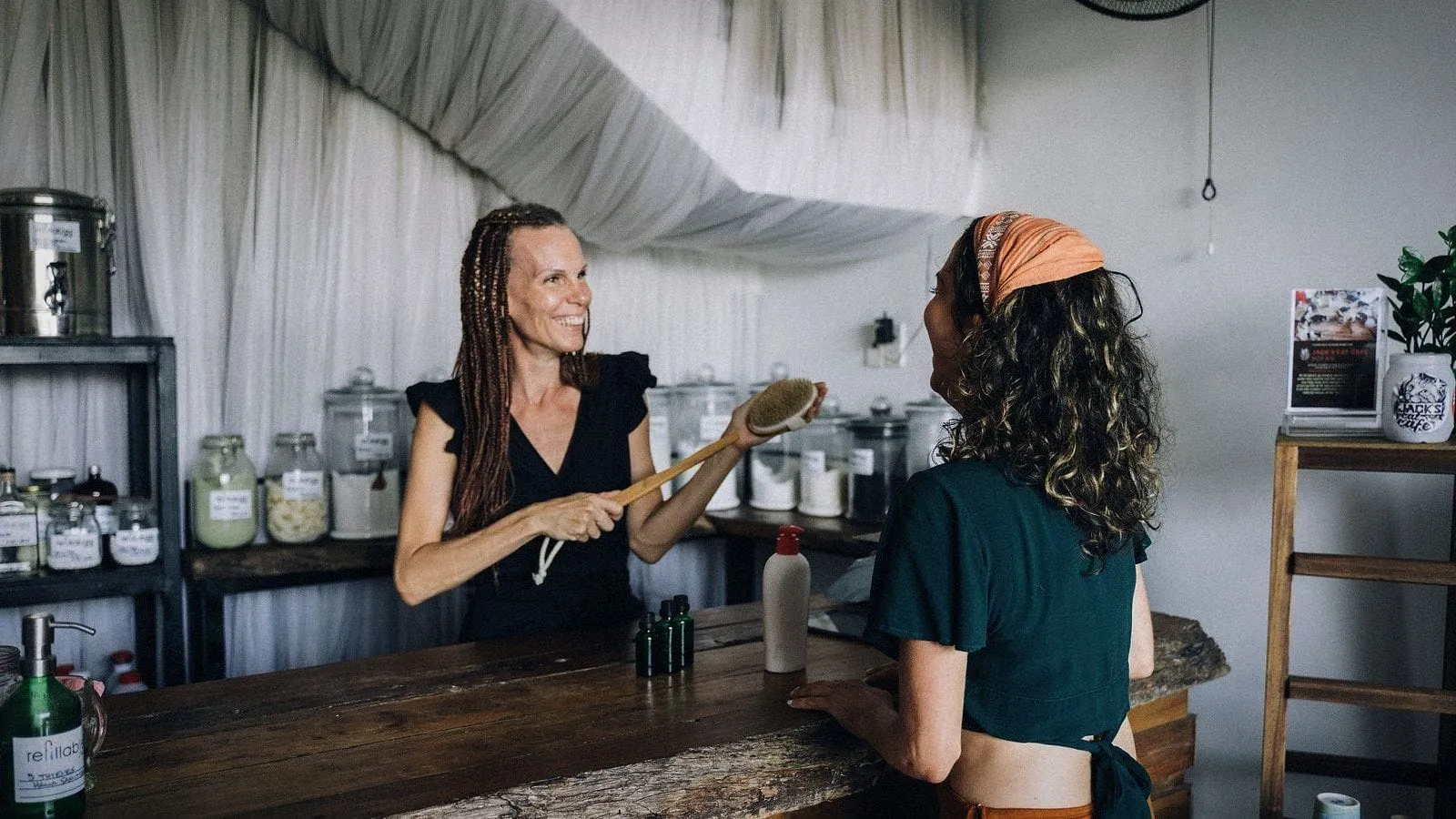 A shop owner laughing with a customer across a wooden counter lined with glass jars.