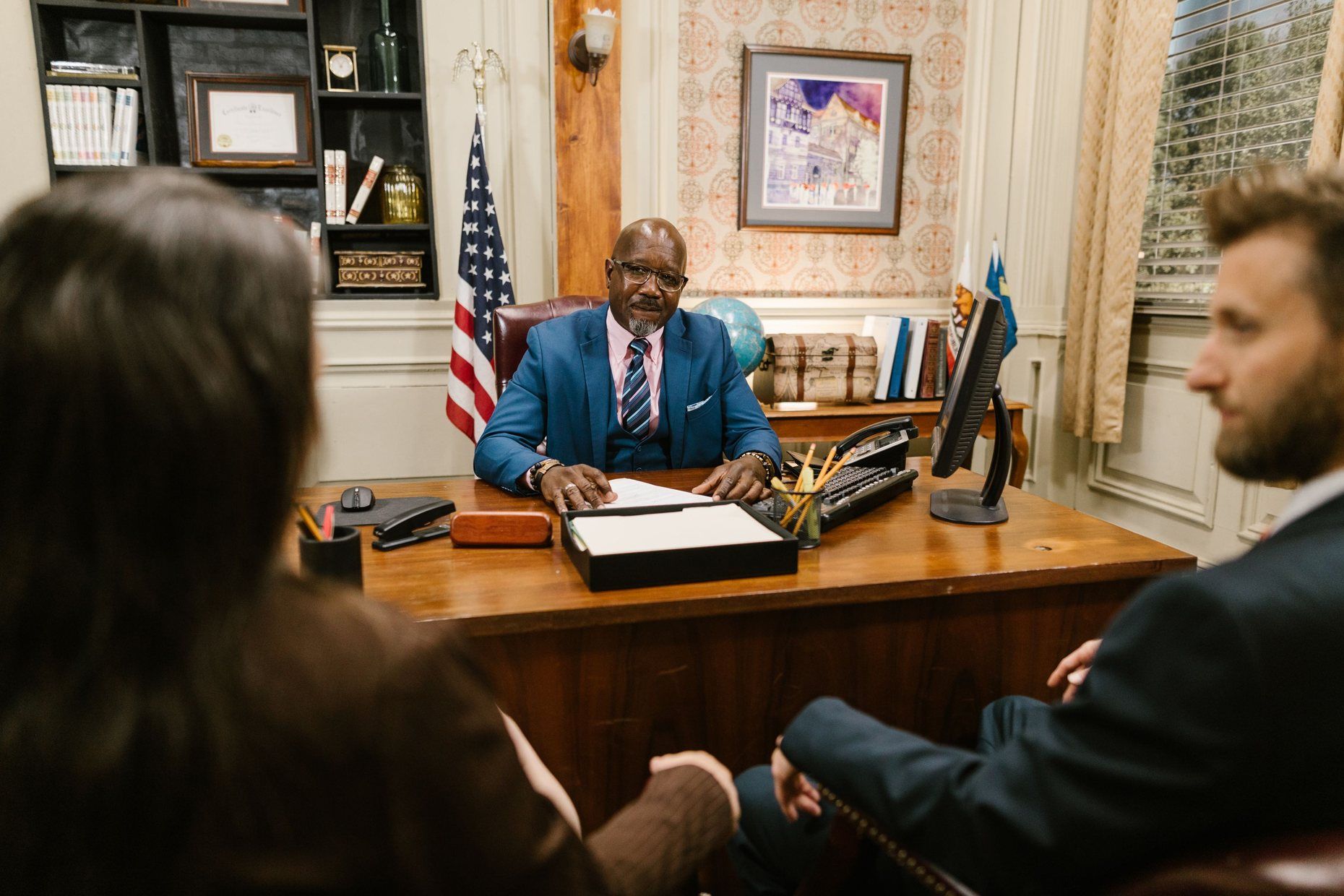 An attorney in a blue suit at his desk, a US flag behind him, listening to two clients seated across from him.