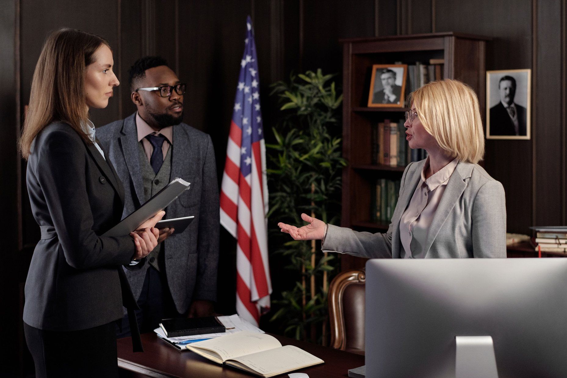 Three lawyers conferring in a wood-panelled law firm office, a US flag and bookshelves behind them.