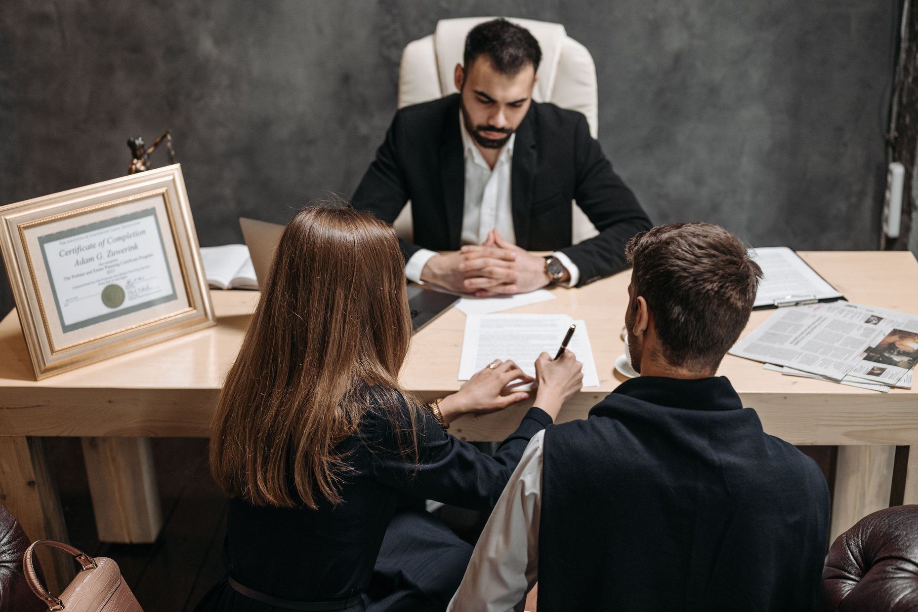 A lawyer at his desk watching two clients sign papers in a small, modern legal practice.