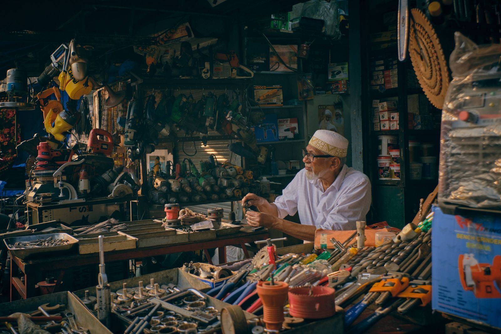 An older shopkeeper sorting tools on the counter of a crowded hardware store.