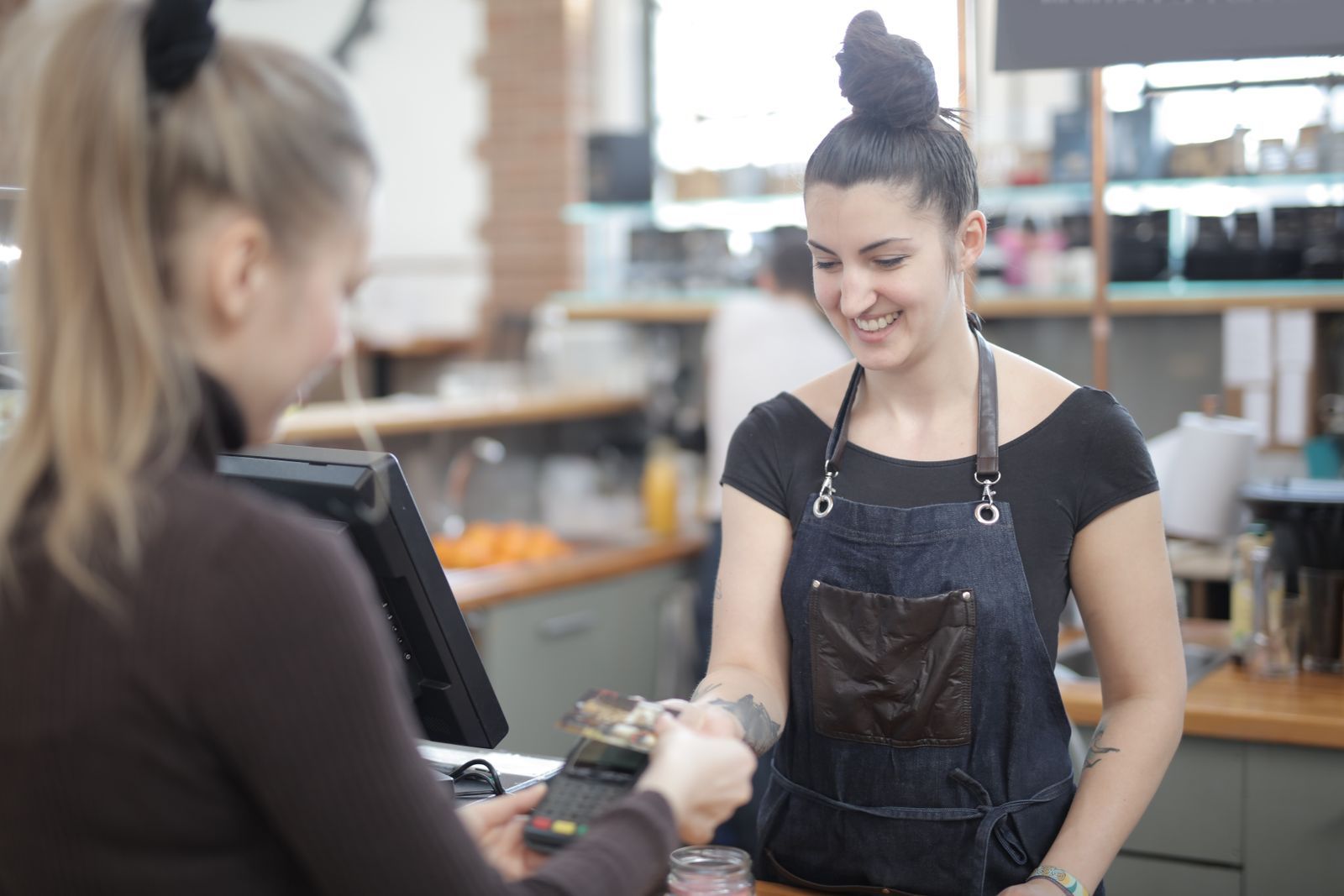 A shopkeeper in an apron smiling as her customer pays at the counter.