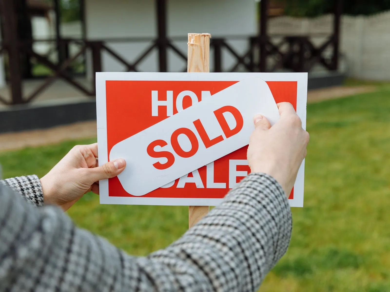 Hands pressing a Sold sticker onto a house-for-sale sign in a front yard.