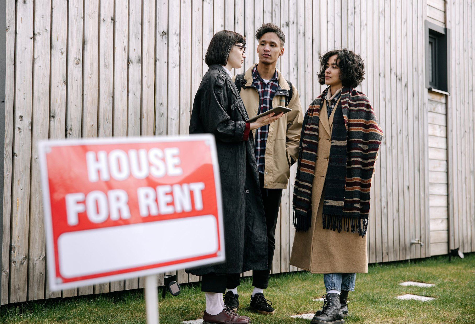 A property manager showing a rental house to two tenants beside a House for rent sign.