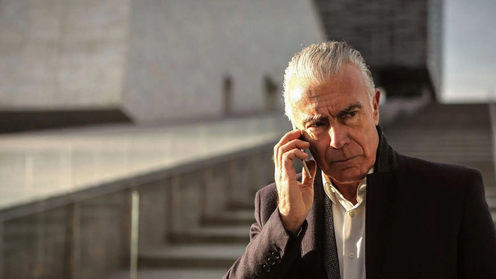 A grey-haired man in a dark jacket on the phone, frowning as he listens.
