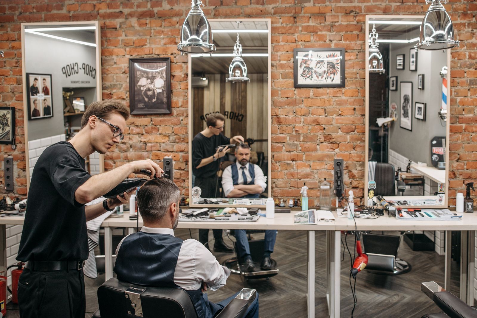 A barber cutting a client’s hair in a brick-walled barbershop.