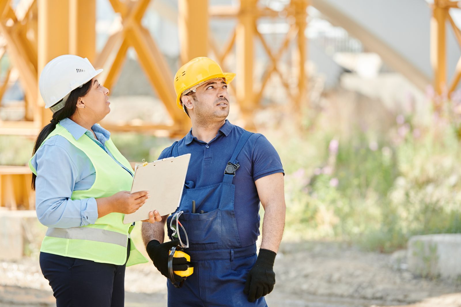 Two site workers in hard hats and high-visibility vests talking on a construction site.