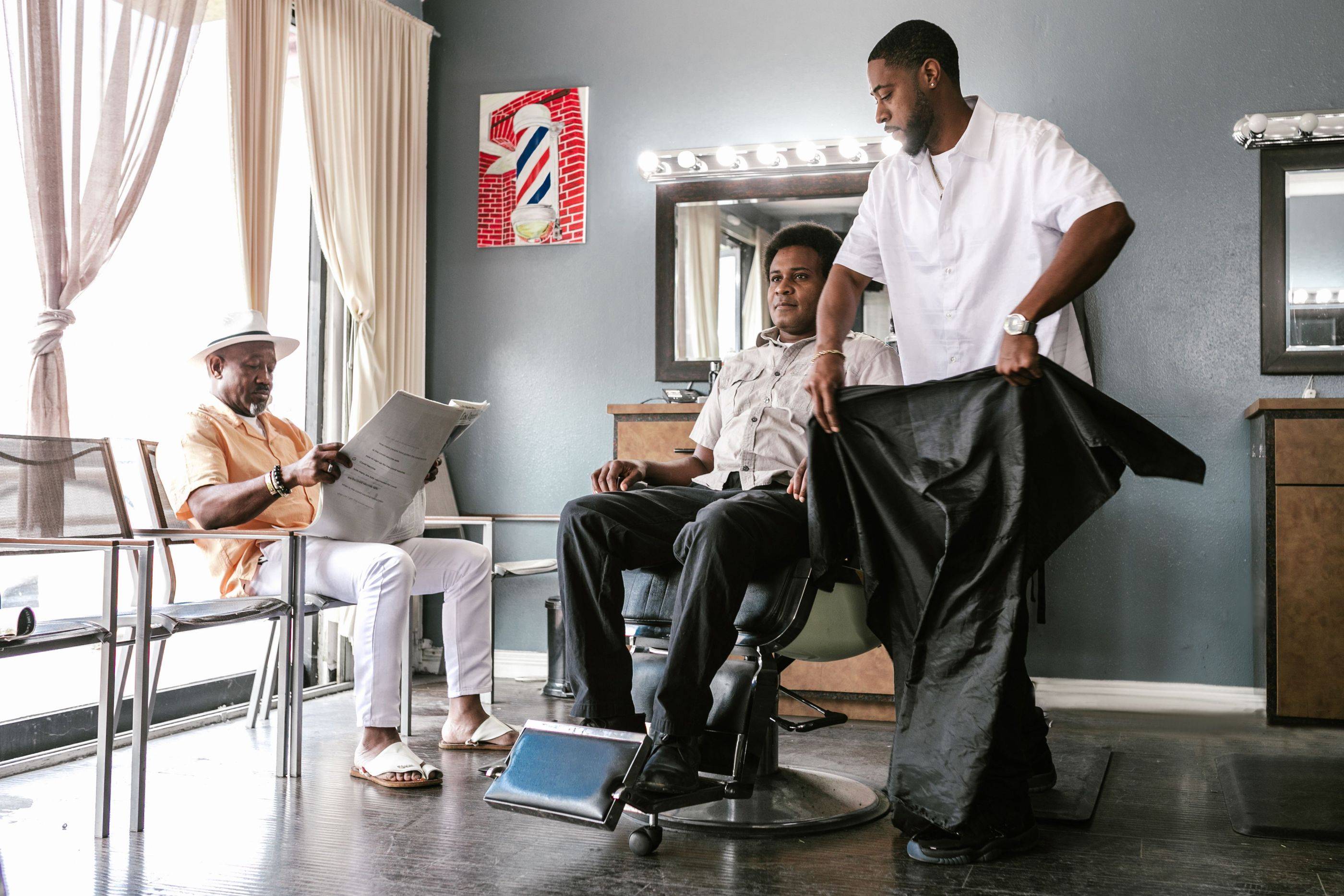 A barber gets a client ready in the chair while the next customer waits with a newspaper