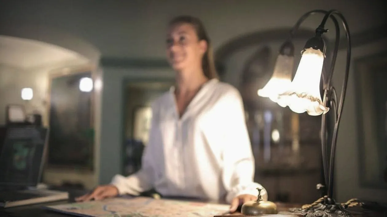 A receptionist at a lamp-lit hotel desk, smiling at a guest.