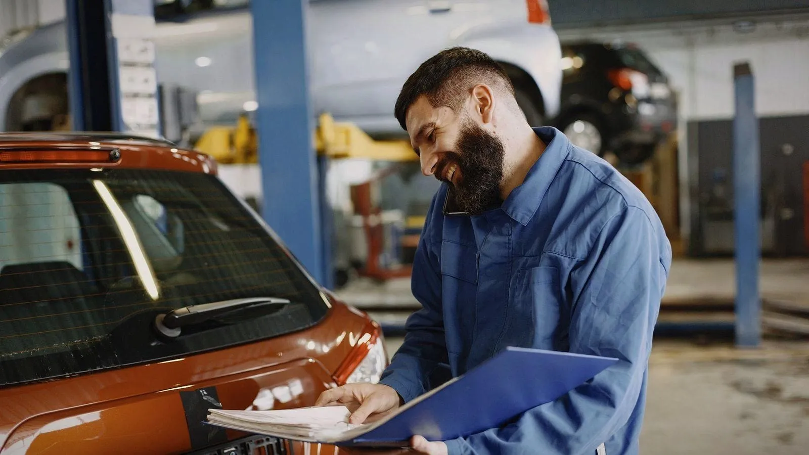 A mechanic in his garage reading the paperwork on a clipboard beside an orange car.