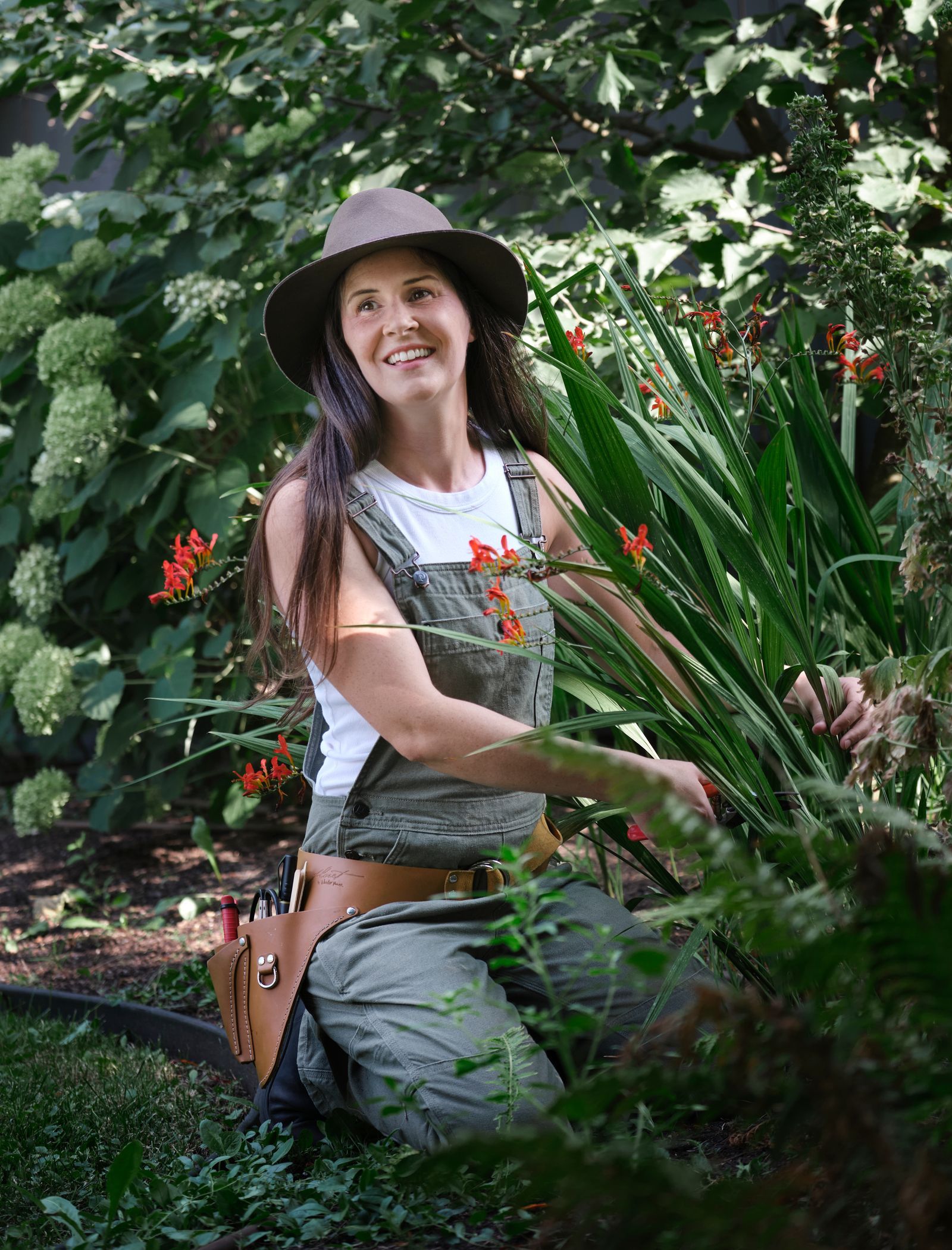 A gardener in overalls and a sun hat tending plants in a leafy garden.