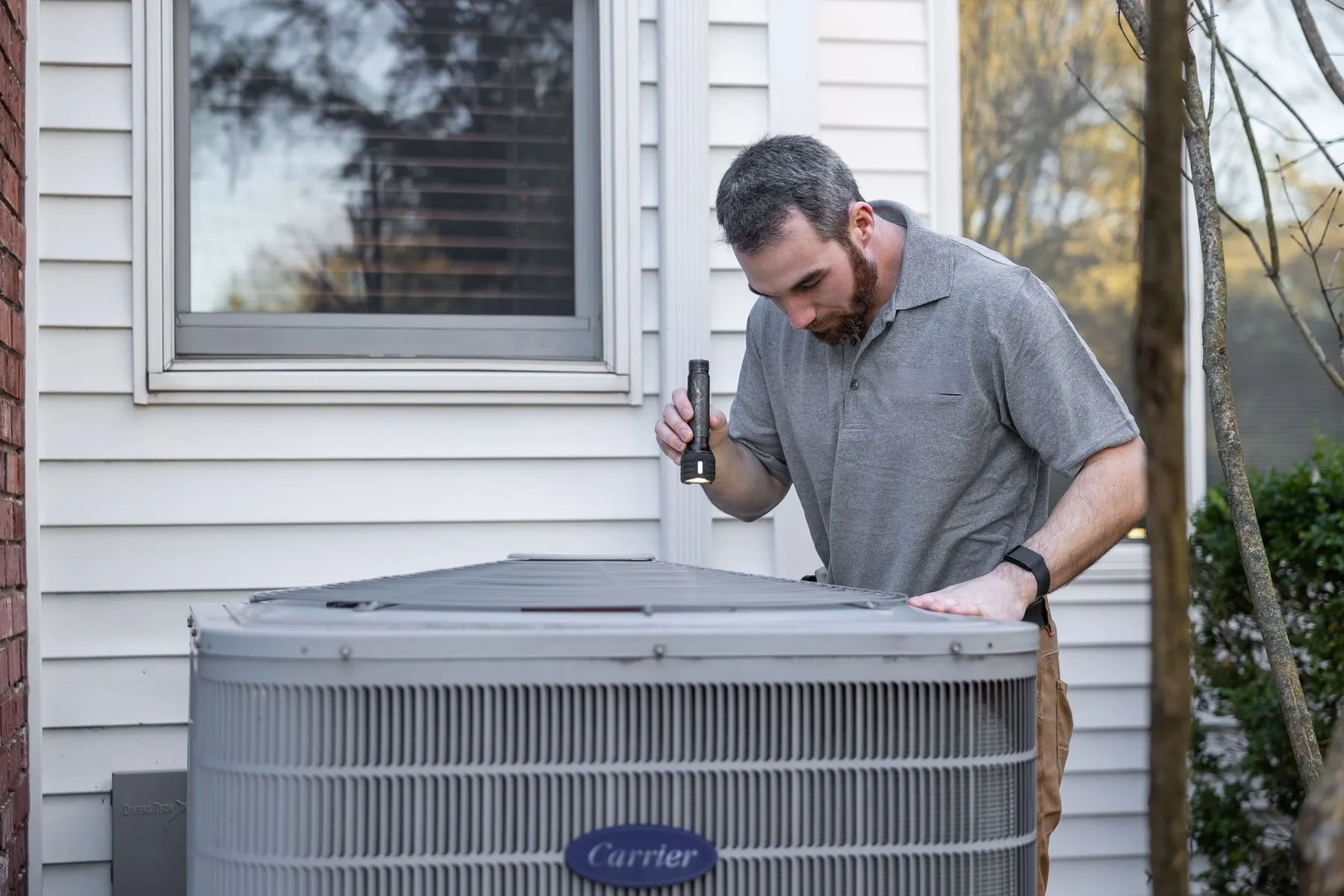 A technician with a flashlight checking an outdoor air-conditioning unit beside a white clapboard house.