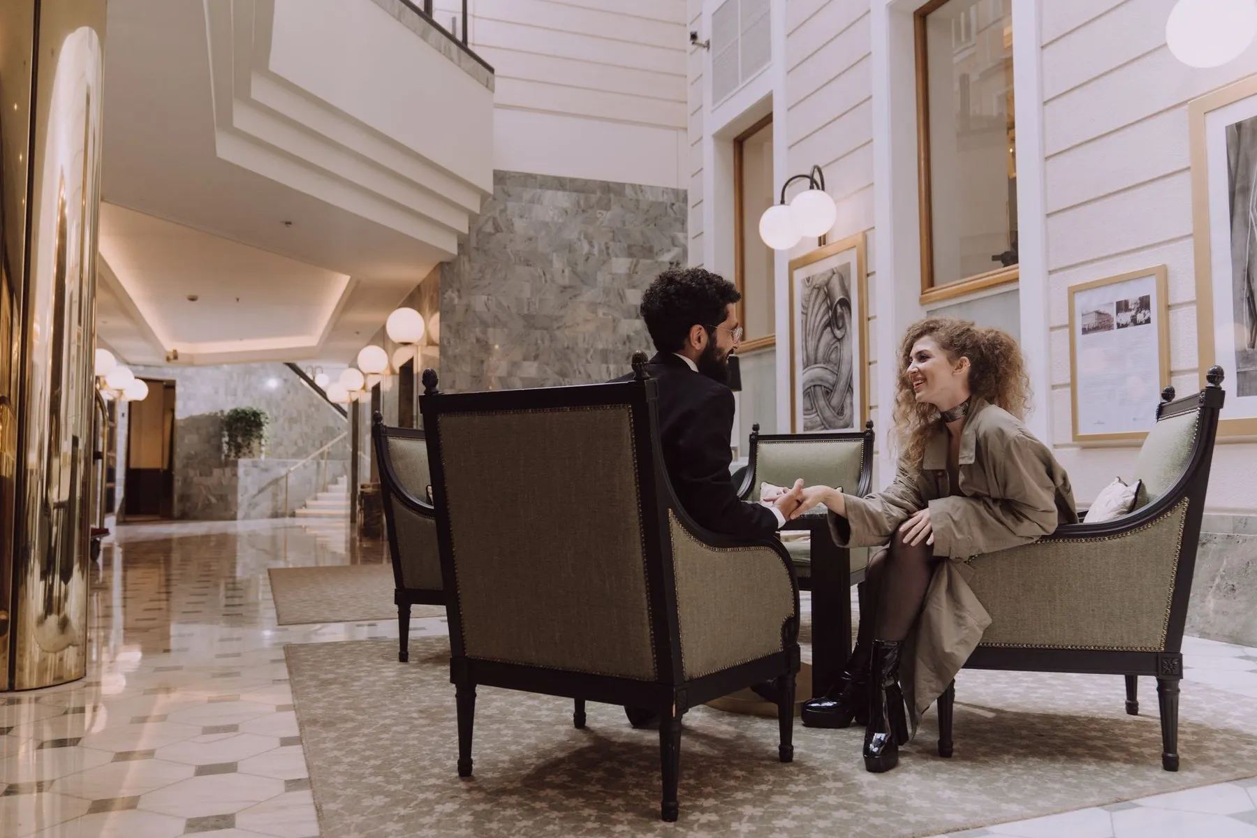 A couple talking and smiling in armchairs in a bright, elegant hotel lobby.