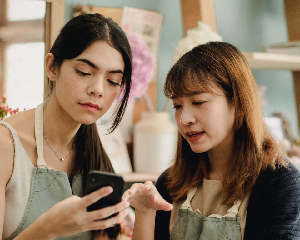 Two florists in aprons stand close together in their shop, reading something on a phone. One points at the screen; both look concerned.