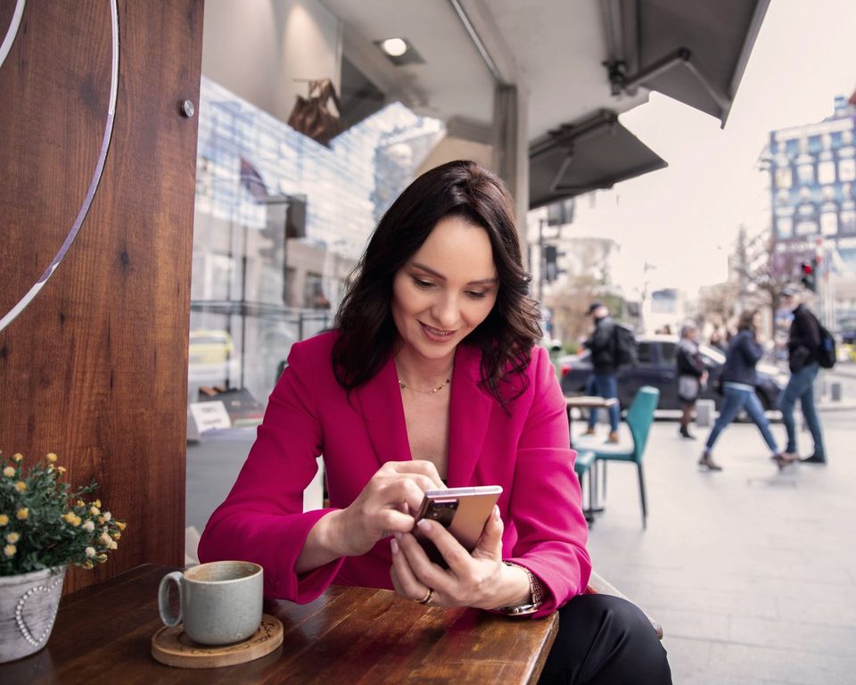 A woman at a café table on a busy shopping street looks at her phone, scrolling, with a cup of coffee beside her.