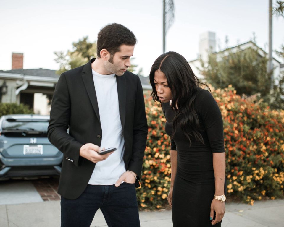 Two neighbors on a residential street lean over one phone. She bends in to read it, taken aback; he scrolls.