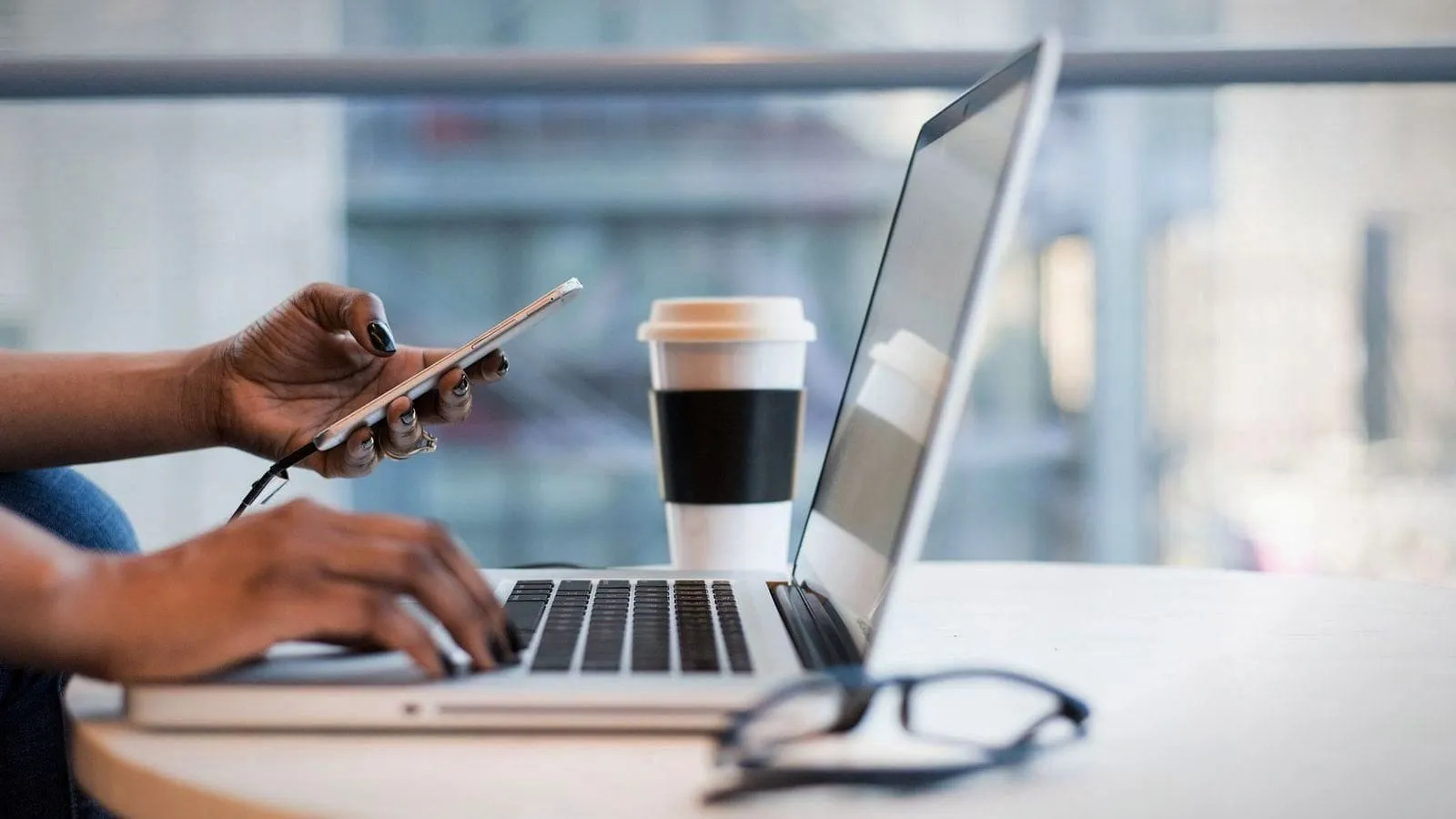 Hands holding a phone and typing on a laptop at a desk by a window, beside a coffee cup.