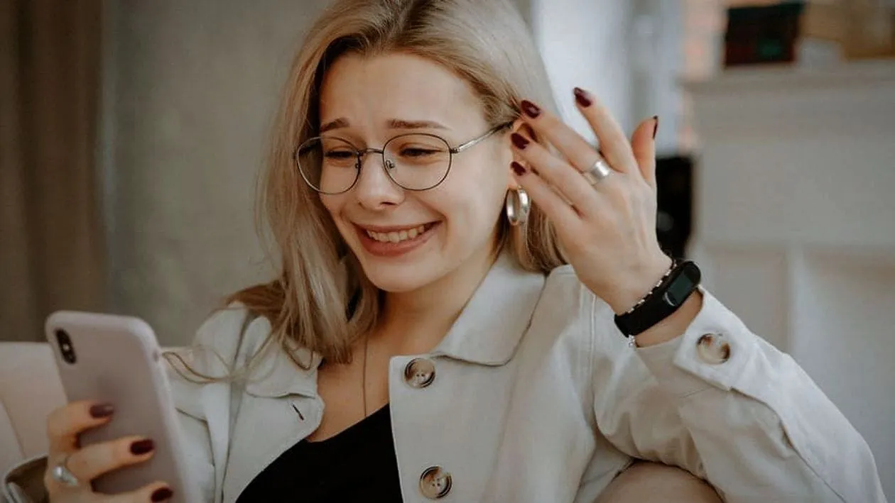 A woman with glasses reading her phone and wincing at what it says.