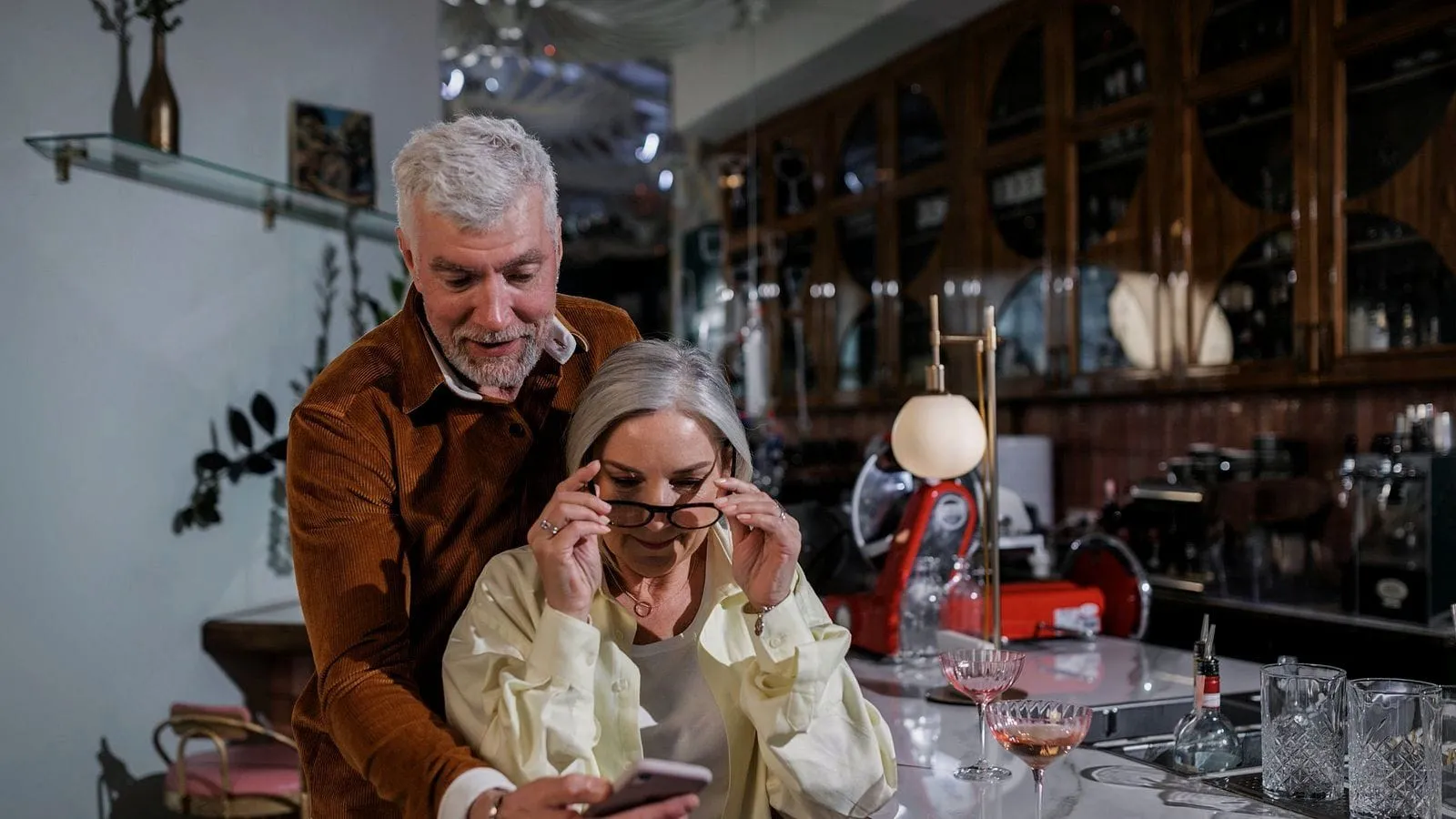 Two café owners behind their bar peering at a phone, she lowering her glasses to read it.