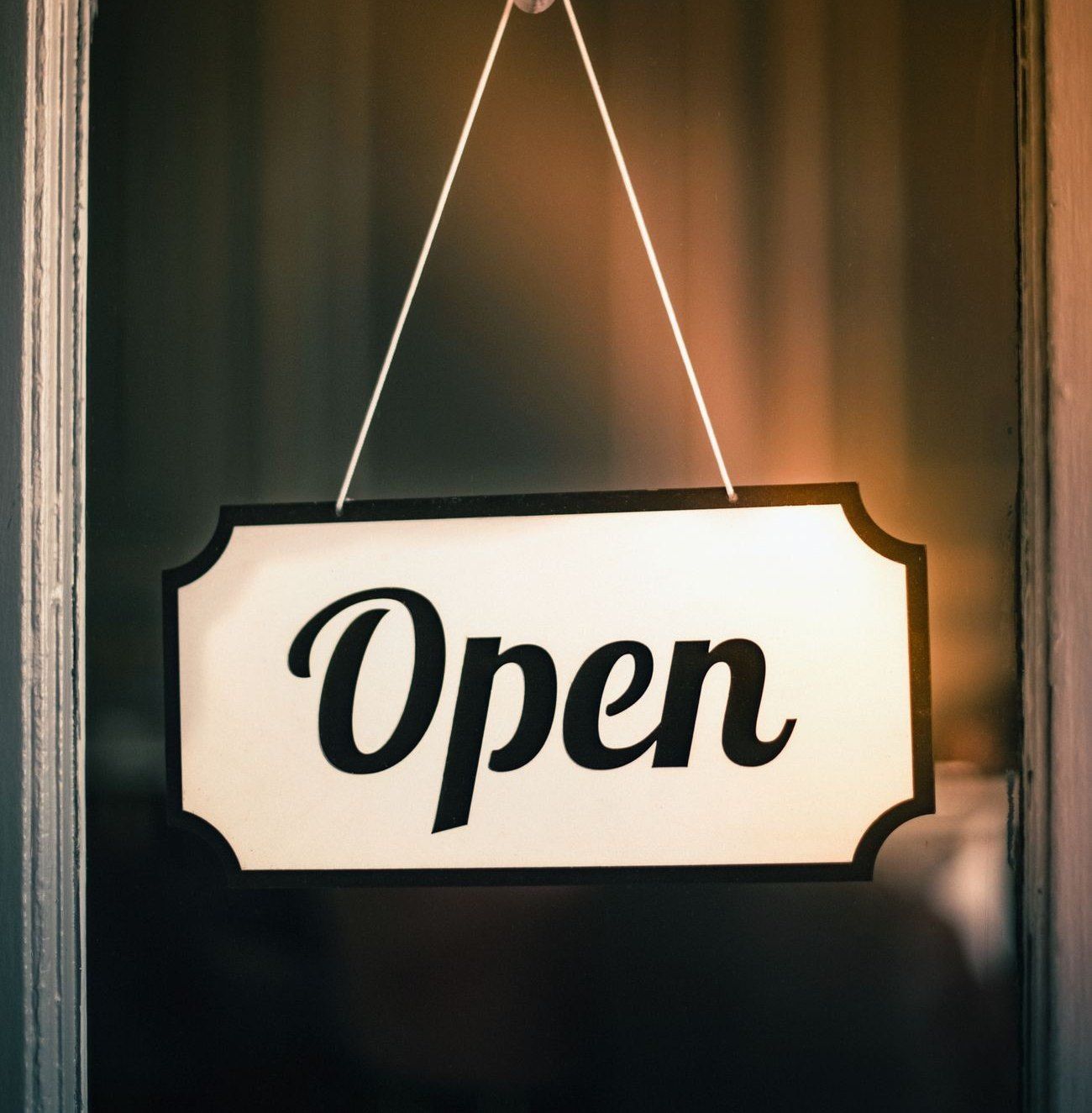 An 'Open' sign hanging on the glass door of a small shop, lit warm from inside.